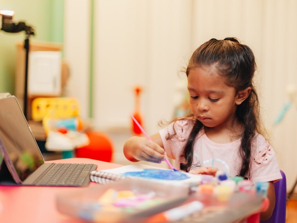 Young girl painting in a notebook while using a laptop, representing creativity and different learning styles in children.