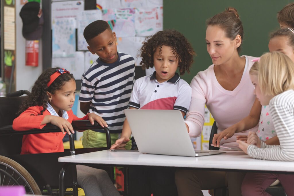 Teacher helping diverse group of elementary students using a laptop in an inclusive classroom environment.