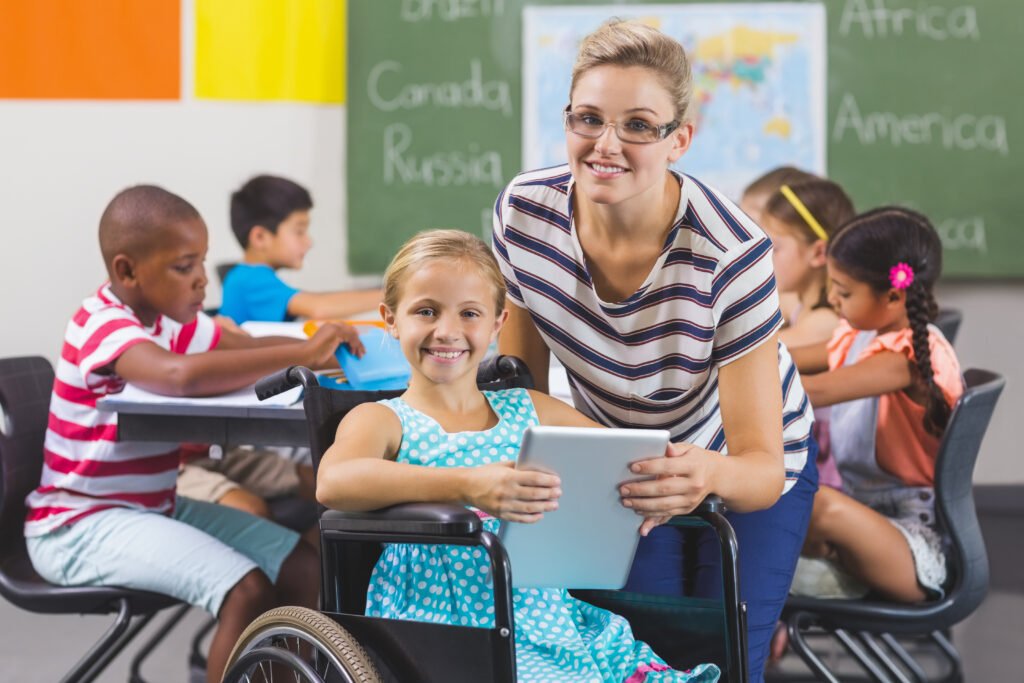 Teacher helping a smiling girl in a wheelchair using a tablet in an inclusive classroom with diverse students.